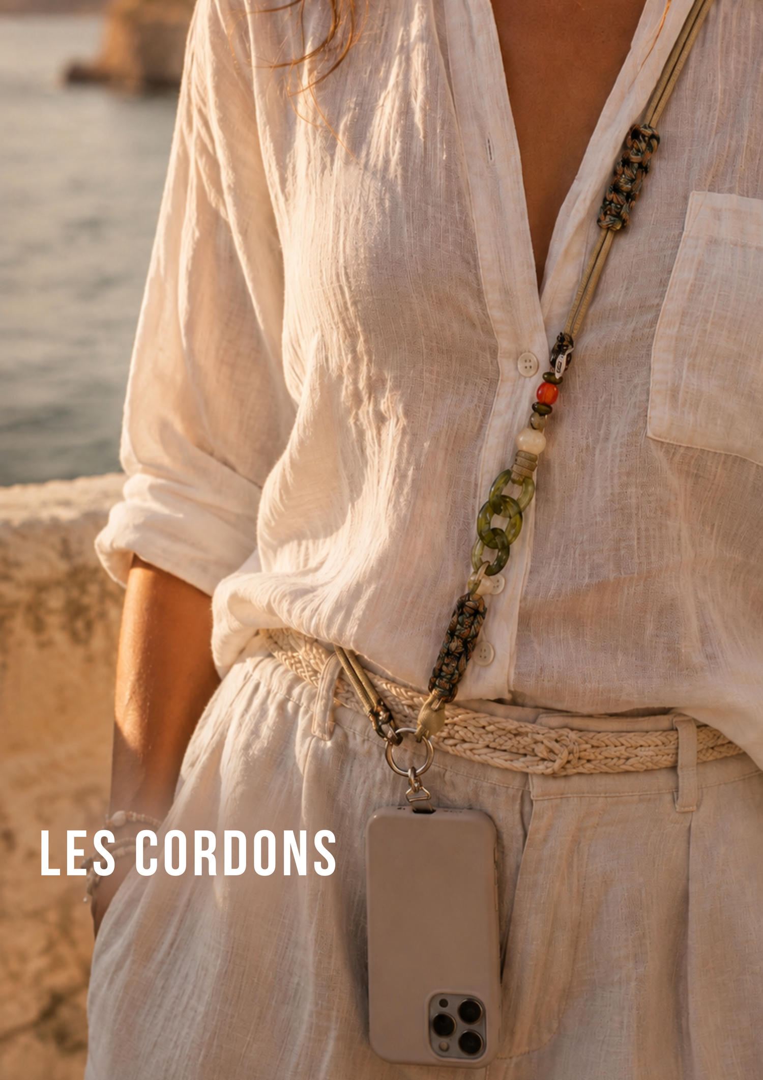 Jeune femme habillée sur une plage avec un bébé sur ses épaules, un téléphone tenu par un cordon de téléphone porté en bandoulière. Les mains de la jeune femme sont donc libres pour tenir son bébé sur ses épaules